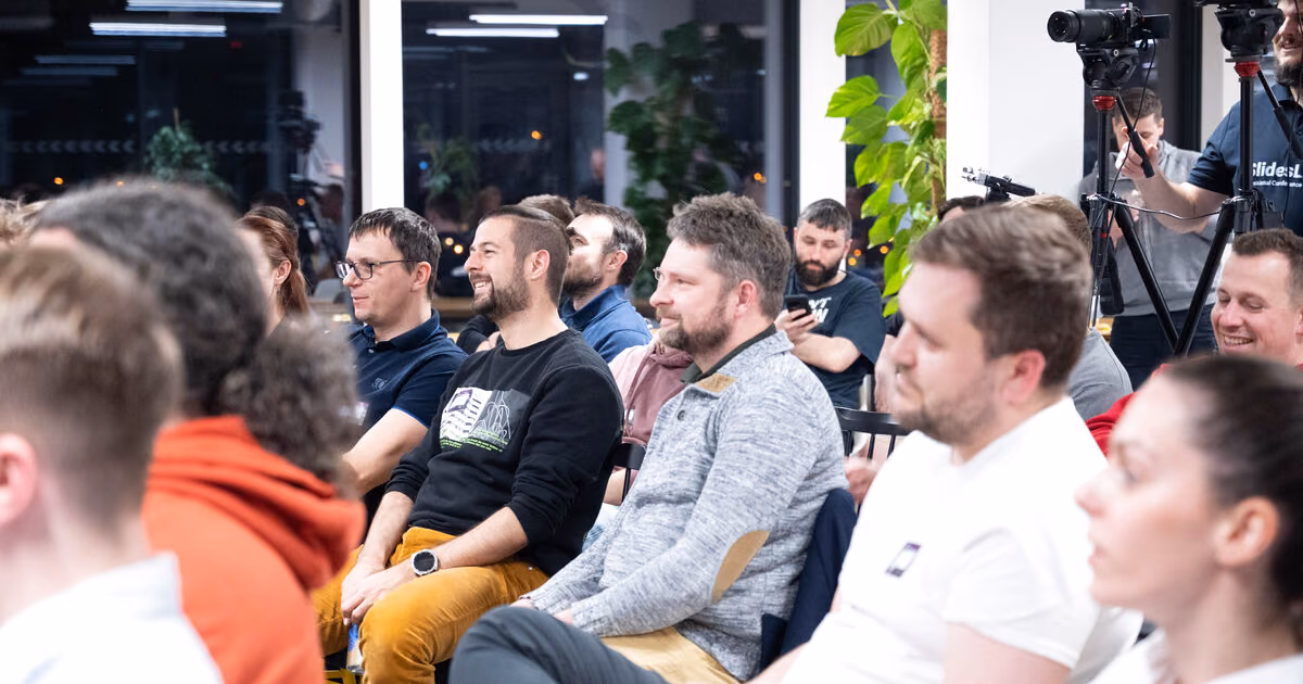 An ELC meetup audience seated near tall windows at night while two videographers film from tripods at the back