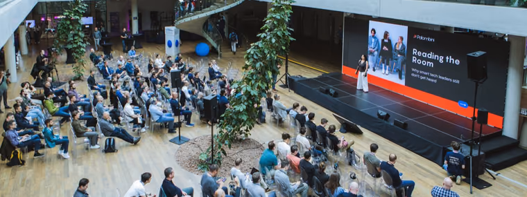 An elevated view of a multi-storey atrium at the ELC Conference, a large seated audience facing a stage with the slide Reading the Room, the cross-functional crowd a Chief AI Officer has to read.