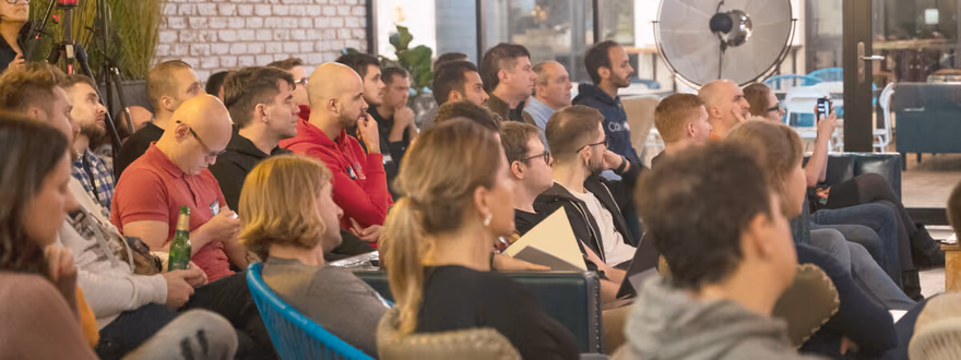 A large group of engineering leaders sitting on lounge chairs and sofas at an ELC meetup in a brick-walled venue lit by pendant lights, listening to a speaker.