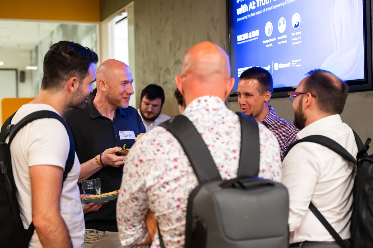 Engineering leadeři s pitím a talíři spolu mluví během networkingové části ELC meetupu, na obrazovce za nimi program večera.