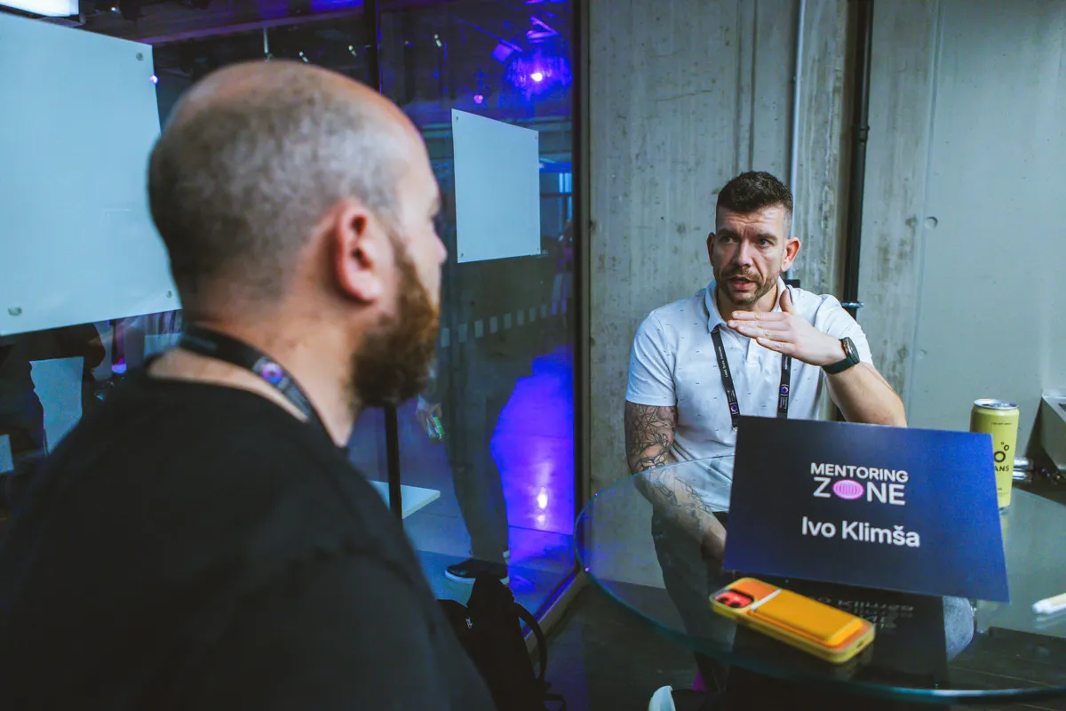 Two people seated at a small glass table under a Mentoring Zone sign at the ELC Conference, one gesturing while talking to the other.