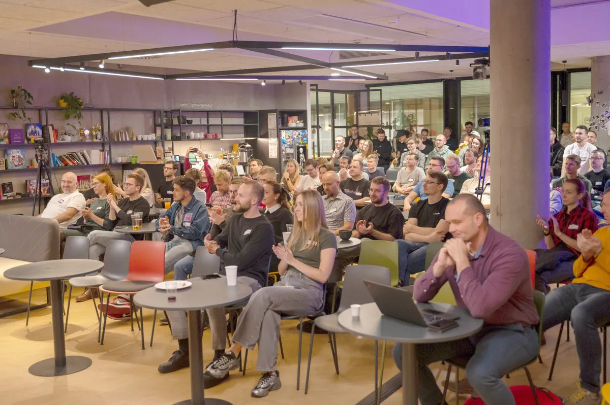 A packed ELC meetup audience seated at small round tables applauding, under string lighting in a venue lined with bookshelves.