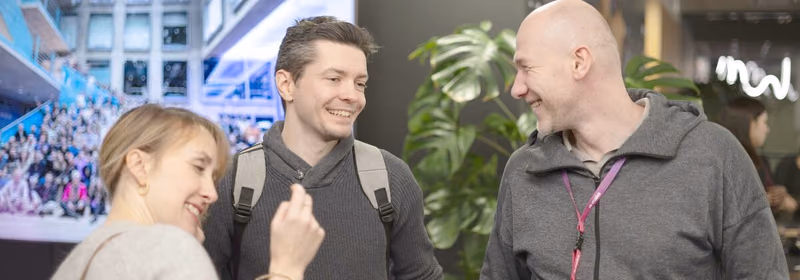 Three ELC meetup attendees standing in a close circle mid conversation and laughing, each holding a drink, one wearing a backpack and an Attendee lanyard, with a blurred crowd photo and a neon community logo on the wall behind them.