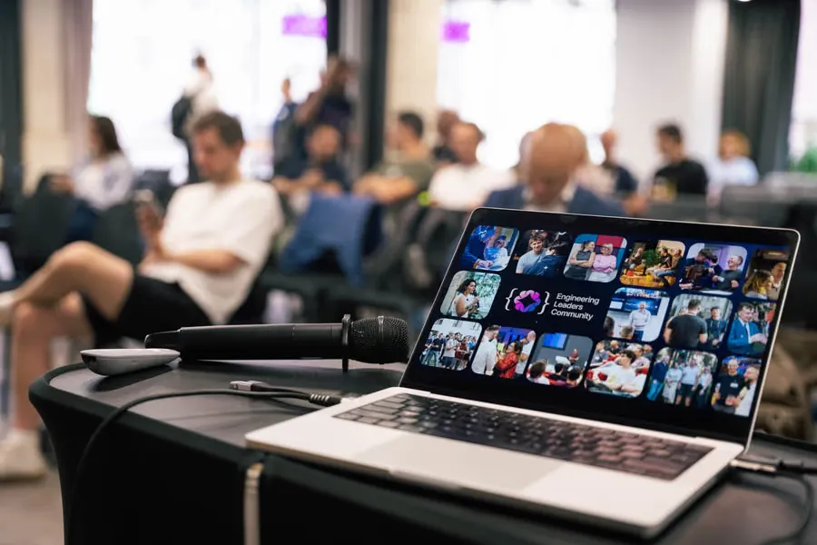A laptop showing the Engineering Leaders Community photo wall, with a full meetup room behind it