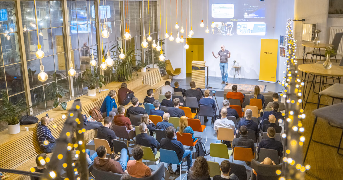 A speaker presenting on a small stage under string lights to a seated audience at an ELC meetup