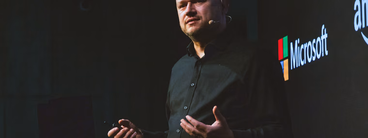 A speaker with a headset microphone gesturing on the ELC Conference stage in front of a screen showing Microsoft, Amazon and other company logos, the vendor landscape a Head of AI chooses from.