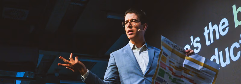 A speaker in a light blue blazer and round glasses wearing a headset microphone on a dark ELC stage, one hand open mid gesture, holding up a large printed sheet headed Feature Roadmap covered in coloured bars, with projected words on the wall behind him.