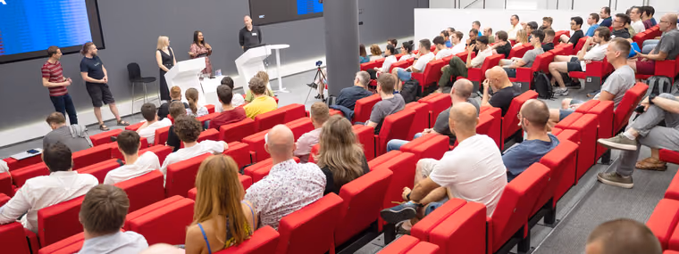 Two women and two men at podiums on a small stage addressing a large seated audience in red auditorium seats at an ELC event, leaders of the org, of teams and of technology on one stage.