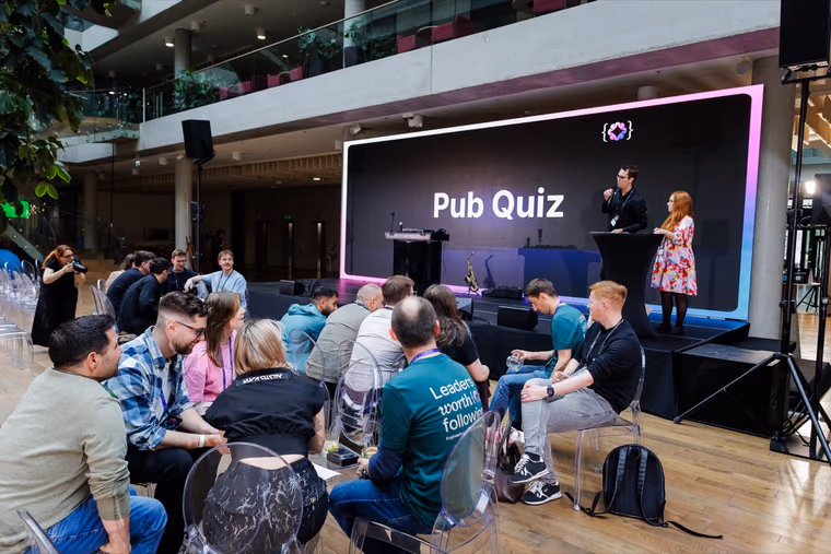 Two hosts on a stage under a large Pub Quiz screen while seated attendees watch, the closing quiz at an ELC event