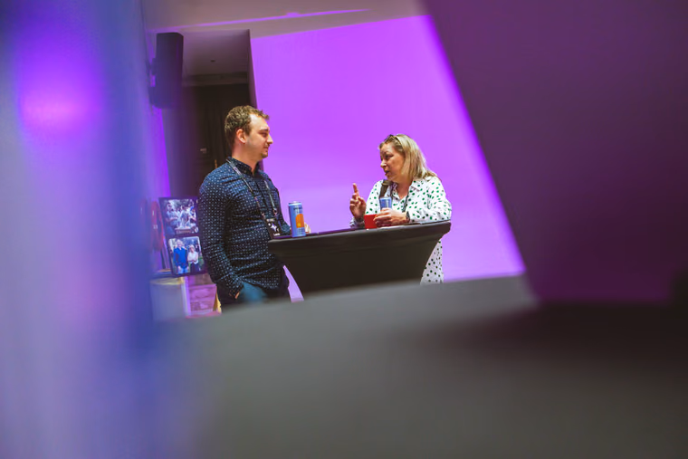 Two attendees in a focused conversation at a standing table under purple light at ELC Conference, the woman making a point with her finger raised