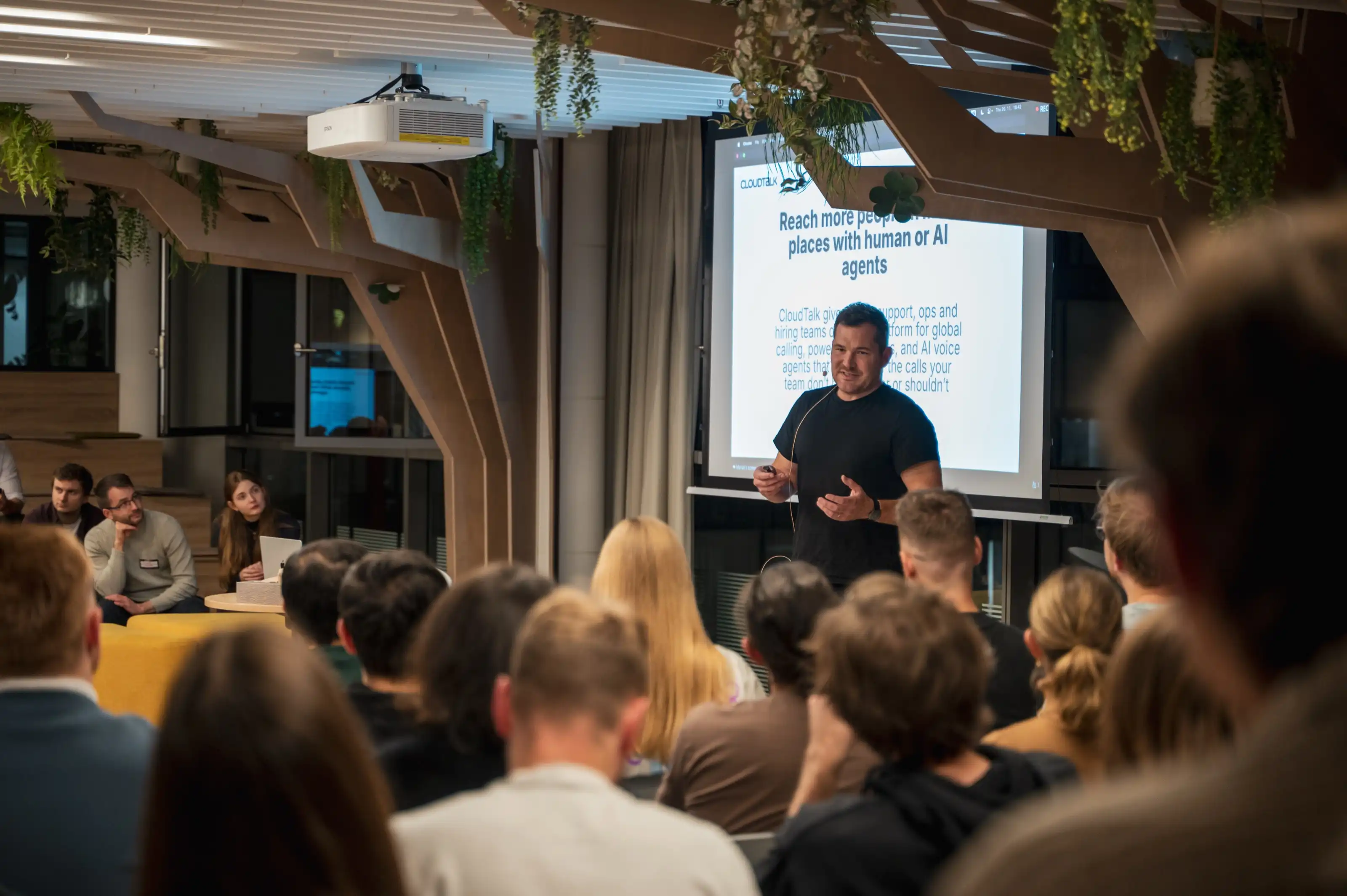 A speaker presenting to a full seated audience beside a product screen at an ELC meetup.