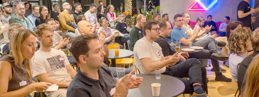 A large seated audience applauding at an ELC meetup, in a room with a lit neon logo on the wall and small round tables holding drinks.