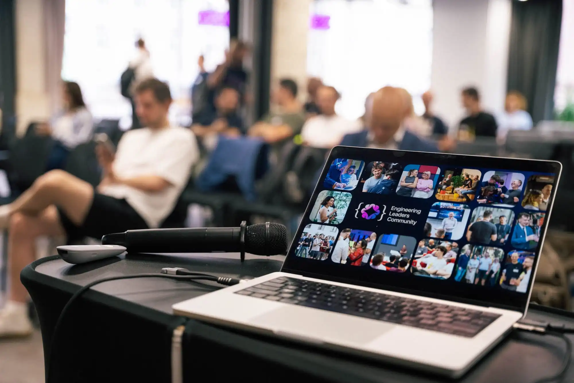 A laptop open to the ELC community photo collage next to a handheld microphone, engineering leaders seated in the audience behind it, the moment just before an ELC talk begins