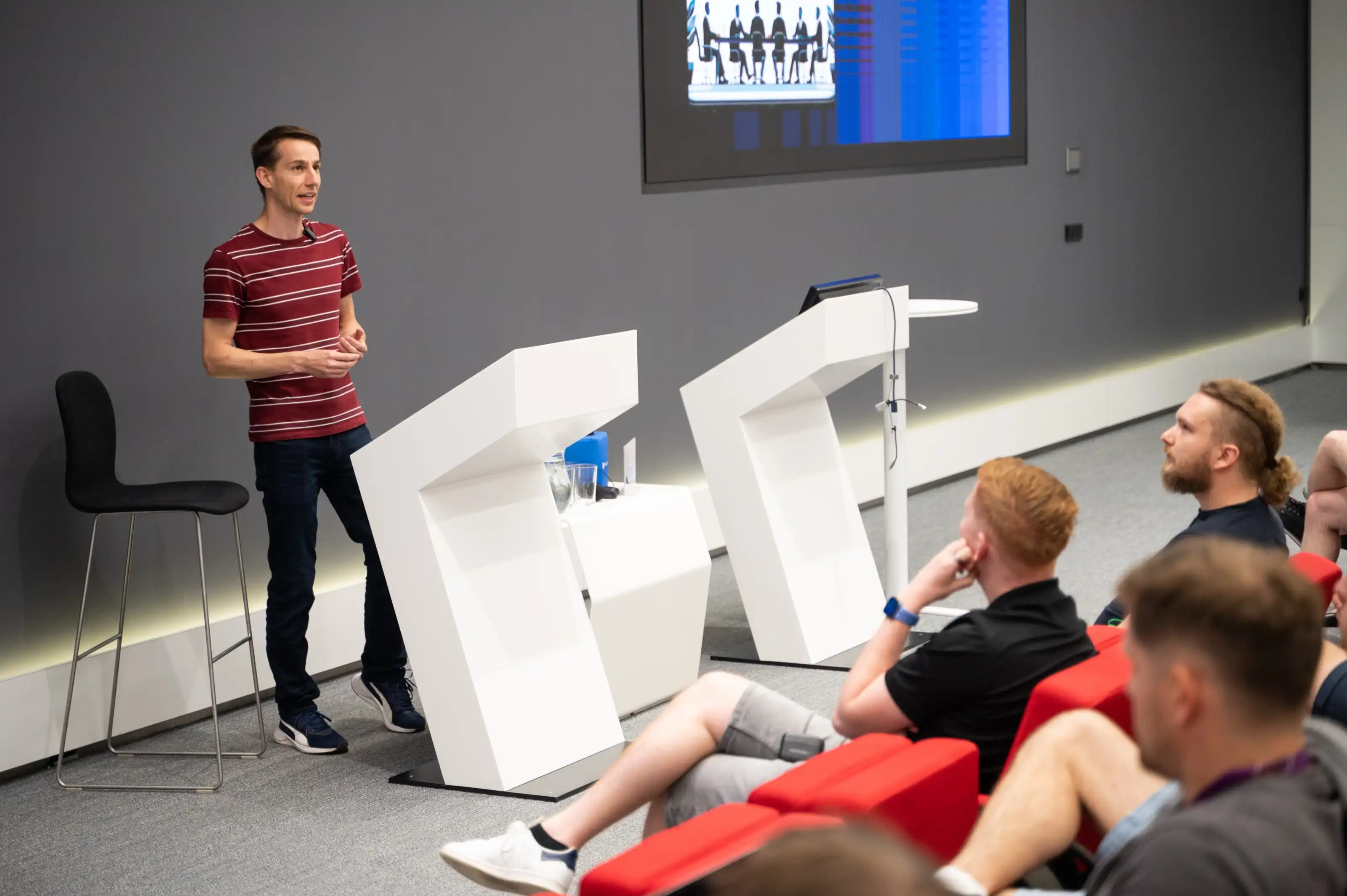 A speaker mid-gesture on stage at an ELC meetup, audience seated in the foreground.