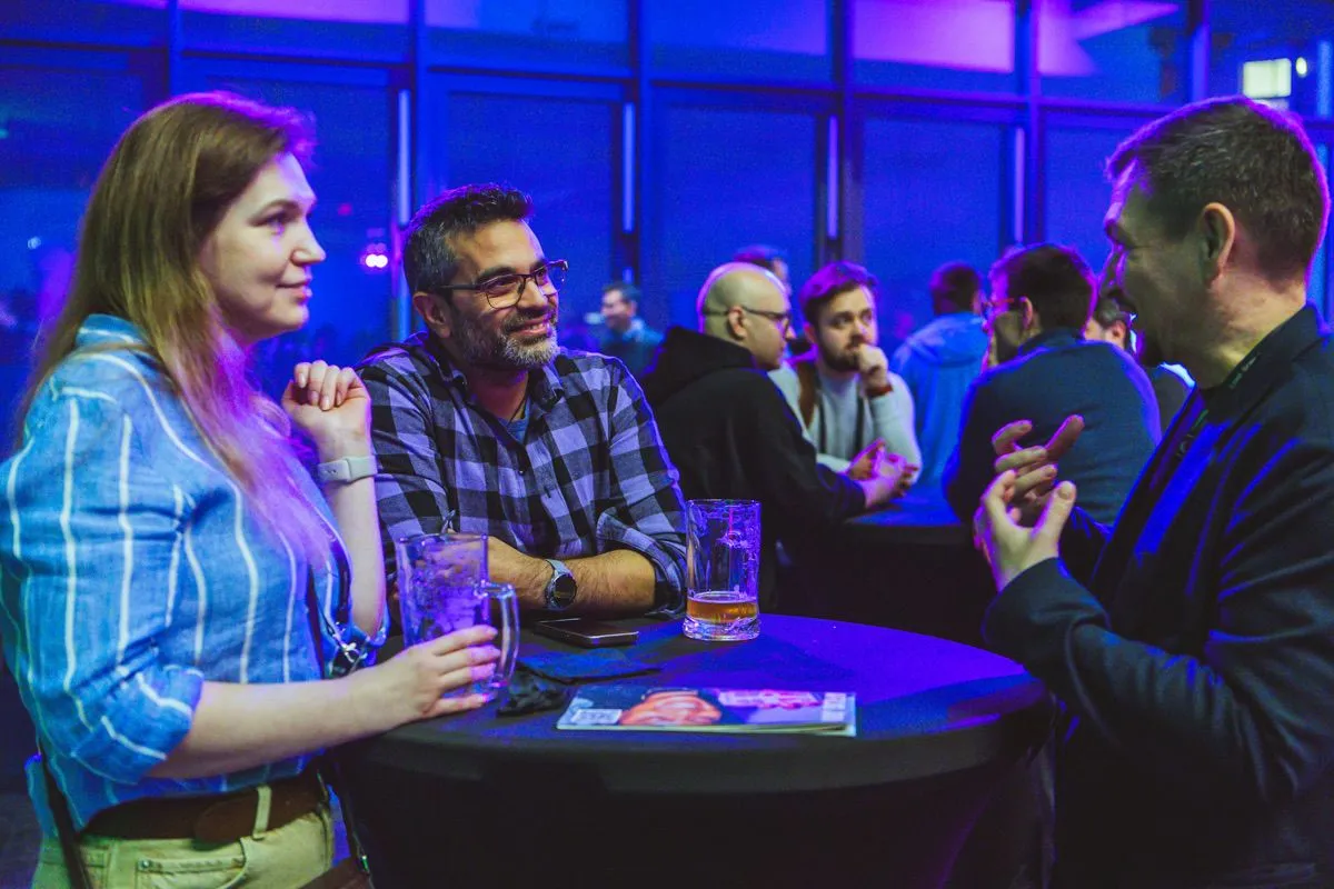 Three attendees talk with drinks at a small round cocktail table in a blue-lit venue while several other guests chat at tables in the background.
