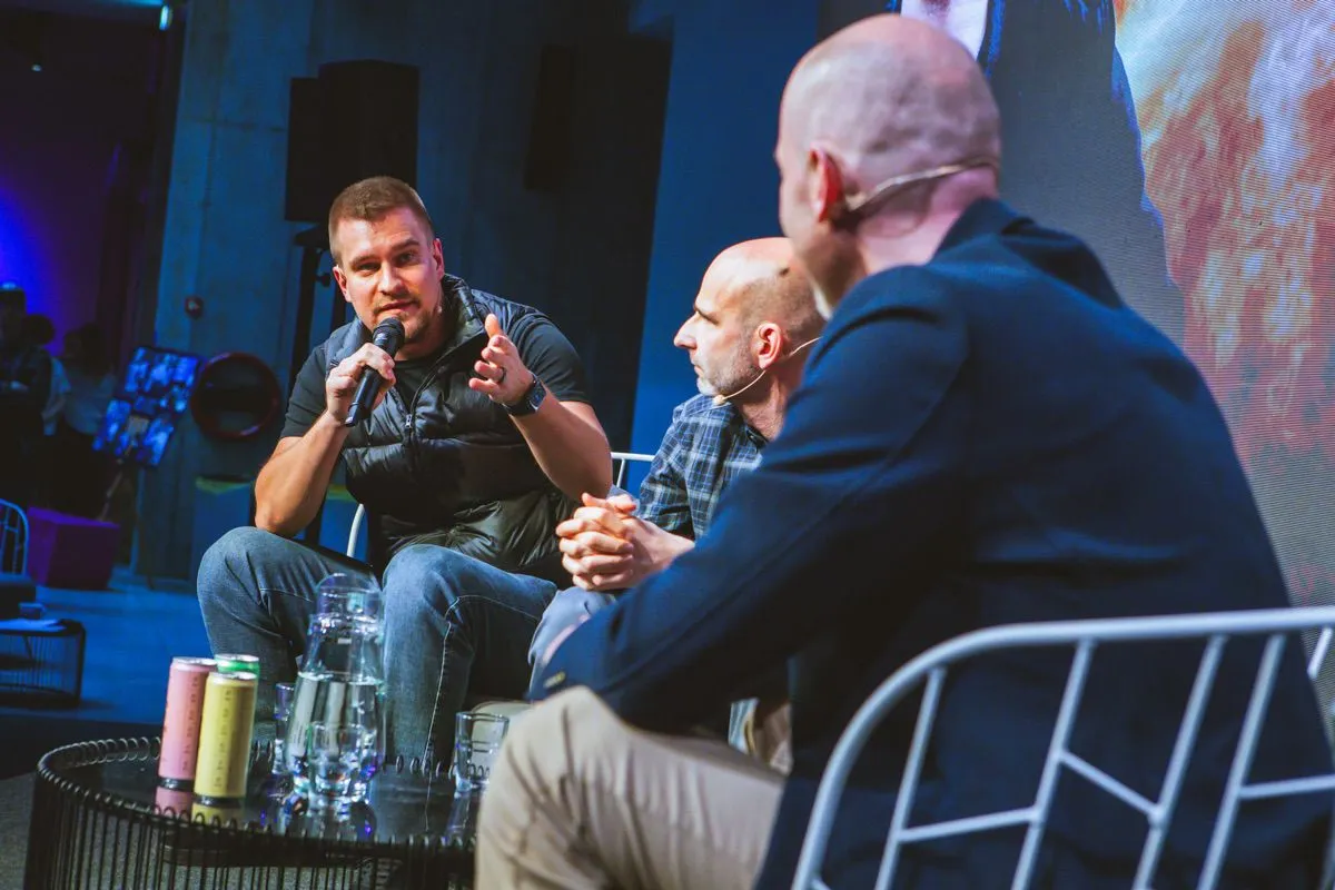 Three panelists sit on a stage during a discussion, with one speaking into a microphone and gesturing while the others listen, and drink cans and glasses on a table in front of them.