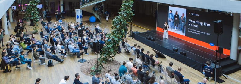 Elevated view of a glass-roofed atrium venue with about sixty attendees seated on clear chairs facing a stage, where a speaker stands in front of a large screen, with a spiral staircase and hanging greenery above.