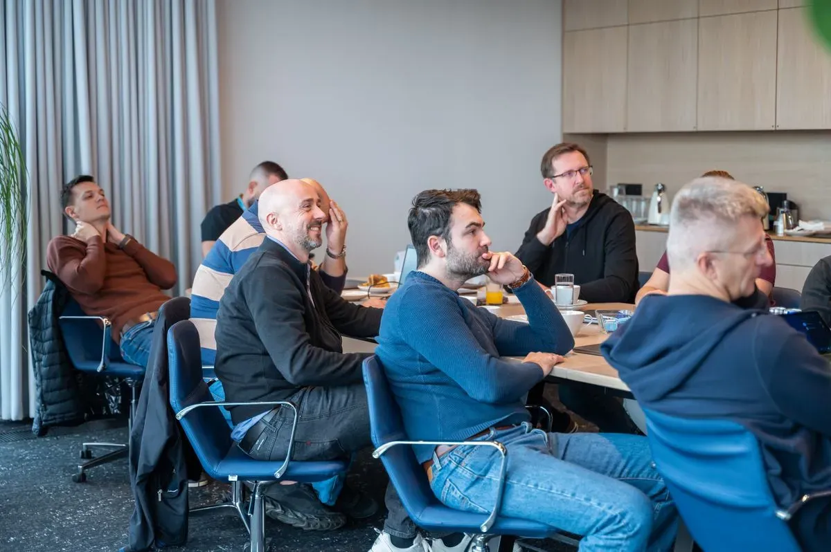 Engineering leaders seated around a workshop table follow the discussion, coffee and laptops in front of them